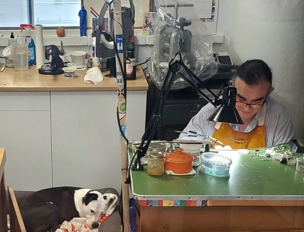 Zachary Androus at his workbench making jewelry with his dog in the background